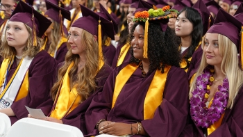 Cronkite School graduates sit in a line wearing their graduation gowns, hats and stoles.
