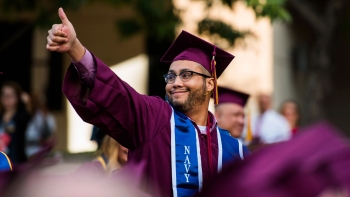 A man in a graduation gown with a Navy military stole smiles and gives a thumbs up to the crowd