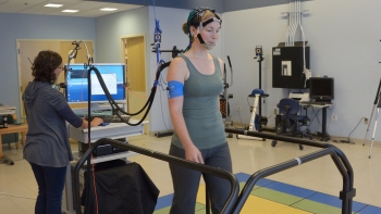A woman with a sensor device on her head walks on a treadmill while a researcher monitors readings on a computer