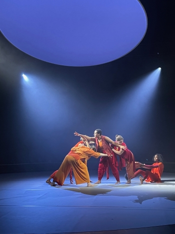 Dancers in bright yellow and red clothing perform on a backlit blue and black stage. 