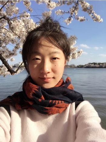 Selfie of Anzi Dong standing in front of a body of water and white cherry blossom tree.