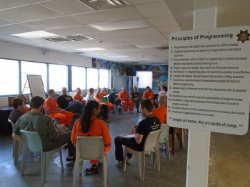Students and people wearing orange jumpsuits sit on chairs in a circle in a prison classroom.