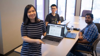 ASU Assistant Professor Jia Zou holding a laptop open as two students at a table behind her look on.