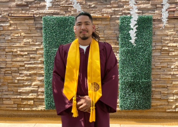 ASU grad Jose Pelagio-Ayala wearing his graduation regalia while standing in front of a brick wall and shrubbery.