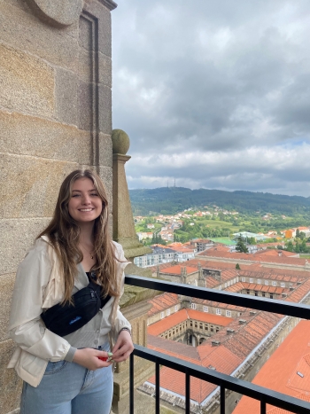 ASU studet Kendall Flynn smiles as she poses on a balcony of a stone building with large buildings with red tile roofs visible in the background below her