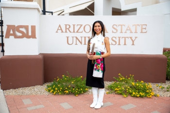 Kristin Payestewa-Picazo stands in front of an Arizona State University sign. She is wearing traditional clothing representing her Hopi and Navajo heritage. She has a feather in between her hands and smiles at the camera.