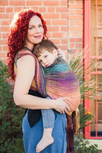 Lela Rankin "babywears" her son in a colorful cloth wrap as both look at the camera, a brick building behind them.