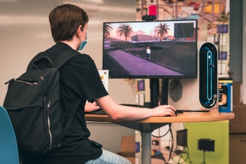 Student with backpack sits at a desk with a computer monitor showing an avatar in a digital twin of the campus.