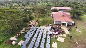 Aerial veiw of a building next to an array of hydropanels in a grassy area. Several people stand near the solar panels.