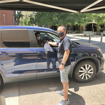 ASU Enterprise Partners CEO Dan Dillon wears shorts, a black t-shirt and black mask and passes out a black t-shirt to employee Peter Means through the passenger window of his blue vehicle. The employee wears a white face mask. 