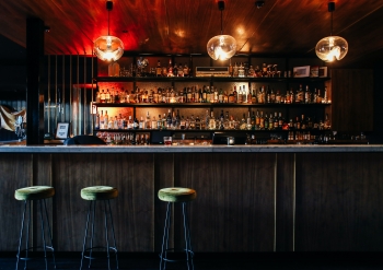 An empty bar with three green barstools in front of it and shelves full of liquor behind it.