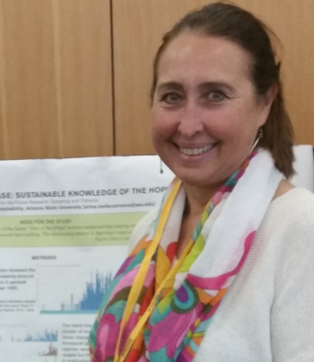 PhD candidate Arina Melkozernova faces the camera and smiles. She is wearing a white shirt with a multicolored scarf and gold lanyard over it. Her brown hair is pulled back behind her ears. Behind her is a poster displaying research.