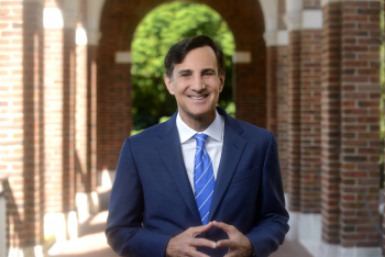 Ronald J. Daniels, president of Johns Hopkins University, is pictured in an outdoor corridor, smiling at the camera with his hands together.