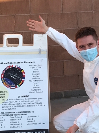 Eli Fox squats in front of a brick wall. He is wearing an all-white jumpsuit with a blue NASA logo over the breast. He has short brown hair and is wearing a blue surgical mask. His arm is extended to point at a sandwich board displaying scientific facts.