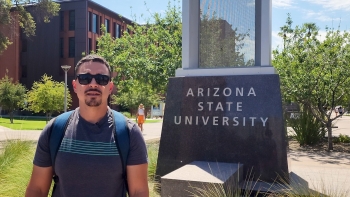 ASU student Gil Ruiz stands in front of a sign on ASU's Tempe campus that reads "Arizona State University."