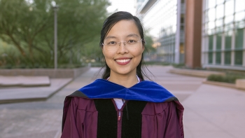 Graduating PhD student Thao Nguyen wearing a graduation robe and standing in front of a building and trees