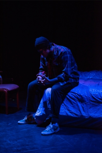 Man seated on a bed on a stage.