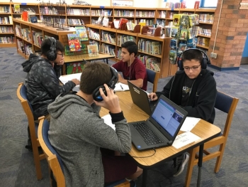 Four middle school students sit at a table in a school library logged in to a digital class, wearing headphones in front of laptops