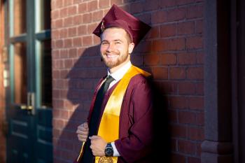 Yusef Sabri leans against the brick side of a building looking at the camera. He is wearing a maroon graduation gown and cap with a gold stole. He is smiling.