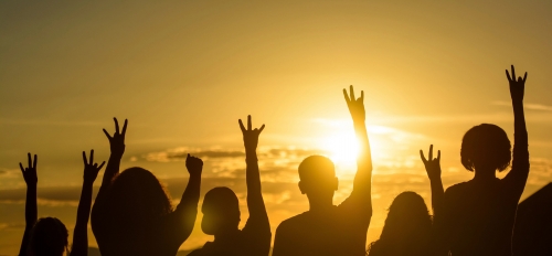 A group of people pictured from the back, silhouetted by a setting sun. They are raising their arms and making the pitchfork gesture with their hands.