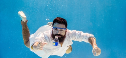 A man in a lab coat is suspended underwater, reaching out for a plastic water bottle in front of him.