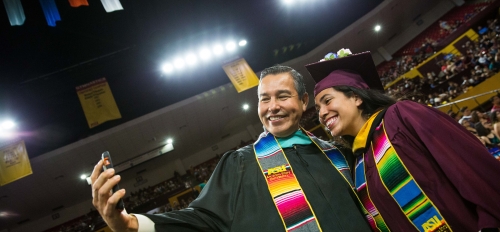 Young student posing with a program director at ASU's Hispanic convocation. Both wear graduation regalia, with the student in a maroon cap and gown and the director in a black gown, and both wear bright, multi-colored stoles.