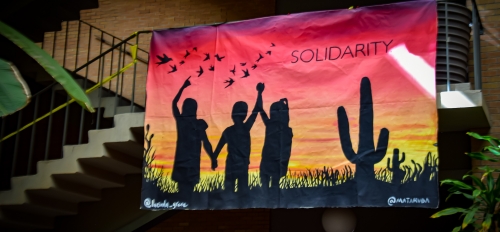 Photo of a mural hung from a staircase featuring silhouttes of three people holding hands in a desert landscape. The mural has the word "solidarity" printed on it.