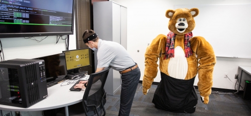 A person works at a computer desk while a large robot made to look like a giant teddy bear stands in the background.
