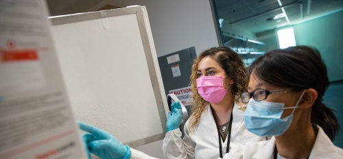 Students in lab coats, medical masks and gloves point at a chart