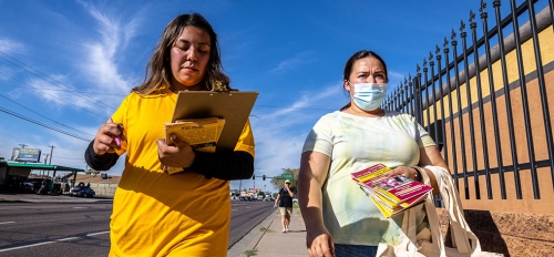 Two women walk down a street holding bags and brochures