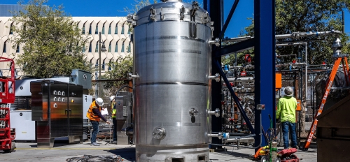 A large metal canister twice as tall as people sits outside surrounded by mechanical equipment 