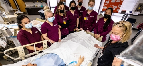 Nursing students look on as an instructor demonstrates at an IV pump next to a manikin. The students are wearing maroon scrubs and surgical masks 