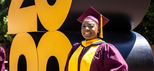 A woman stands proudly in graduation cap and gown in front of a giant 2022 sign