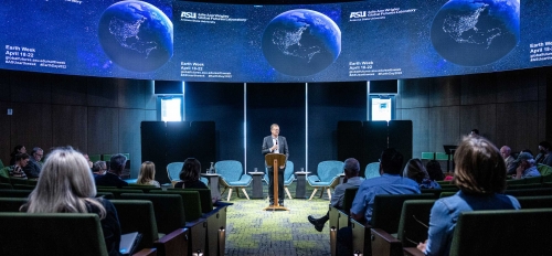 Man speaking behind a lectern at an event with TV screens displaying Earth behind him.