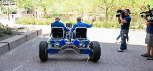Two men in a lunar rover drive away on a sidewalk.