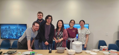Six individuals, students and faculty of the Japanese department smiling at the camera in front of a table with bowls of rice, a rice cooker and seasoning placed on the table.