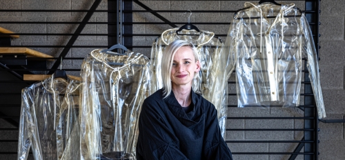 A woman with blonde hair wearing all black sits in a chair with see-through raincoats made of algae polymer hanging on a rack on the wall behind her.