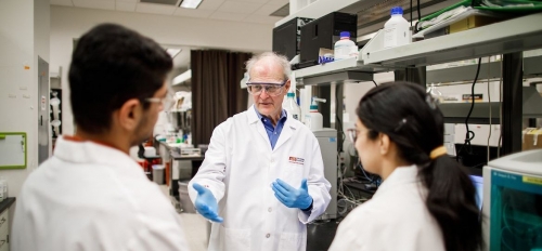 Bruce Rittmann, wearing a white lab coat and goggles, talks to two fellow researchers. 