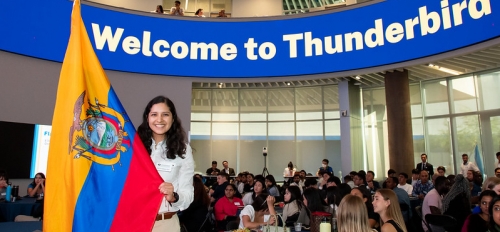 Patricia Baez presenting the flag of Ecuador in front of a large blue and white sign reading "Welcome to Thunderbird"