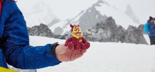 Hand holding a stuffed Sparky the Sun Devil against a backdrop of a snowy, mountainous landscape.