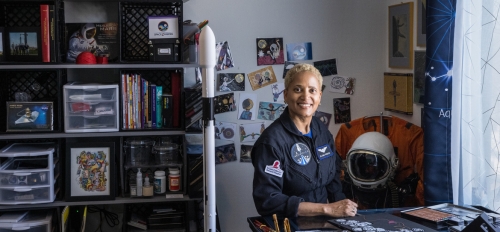 Smiling woman in a NASA space suit sitting in an office surrounded by space-themed paraphernalia.