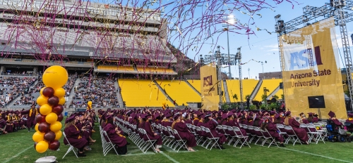 Graduates in caps and gowns seated in folding chairs on a football field as maroon and gold streamers fly overhead.