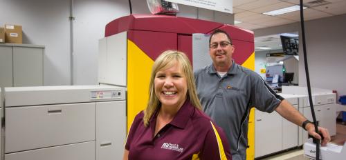 two ASU staff members posing in front of large printer