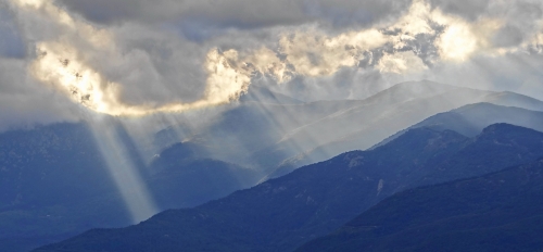 Light rays shining through clouds onto a mountainous landscape.