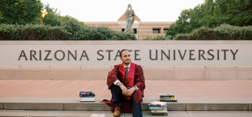 Bryson Brown sitting on steps in front of Arizona State University in graduation gown surrounded by books