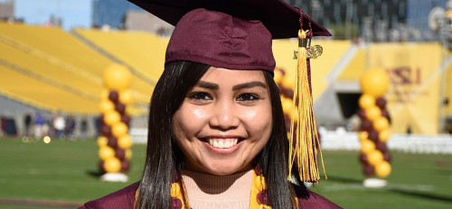 Danni Baquing smiling and posing with ASU degree wearing graduation gown and stole in an outdoor setting with maroon and gold balloons behind her.