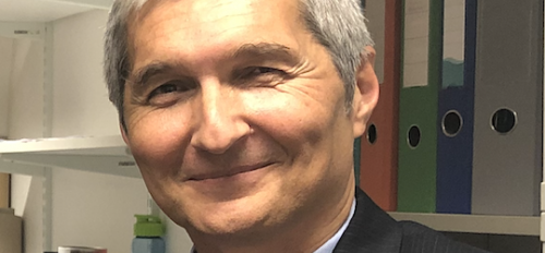 ASU Professor Danko Šipka stands in his office on campus. He has silvery hair and broad smile, but isn't showing teeth. He is wearing a pale blue dress shirt, red tie and black suit coat.