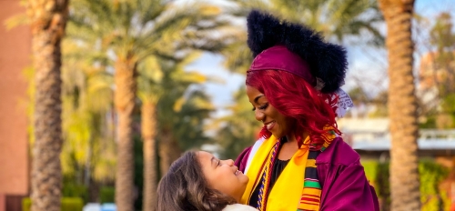 ASU grad Edeline Plaisival in her cap and gown on Palm Walk smiling at her 8-year-old daughter