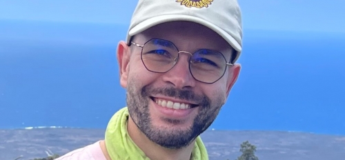 Florian smiling at the camera wearing an ASU hat, a pink shirt and a green neck gaiter with an ocean in the background