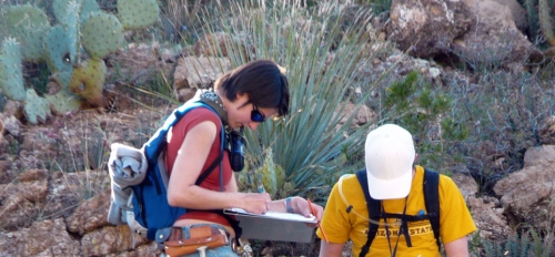 Two students in jeans and T-shirts taking notes outside in a desert environment.